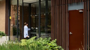 Un edificio moderno con entrada de vidrio, una persona caminando adelante y una puerta con cámara de seguridad.