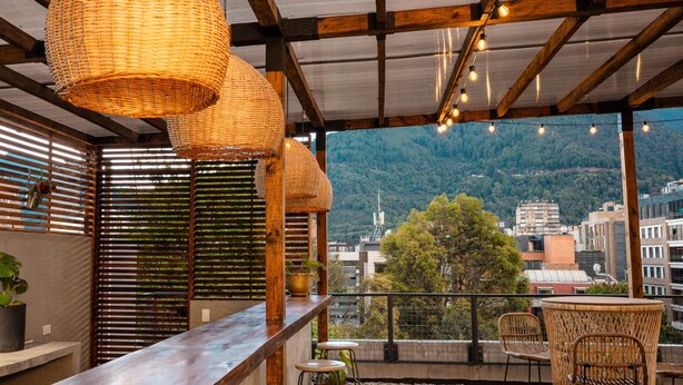 Terraza de madera con mobiliario de mimbre, zona de bar y vistas a una ciudad y montañas.