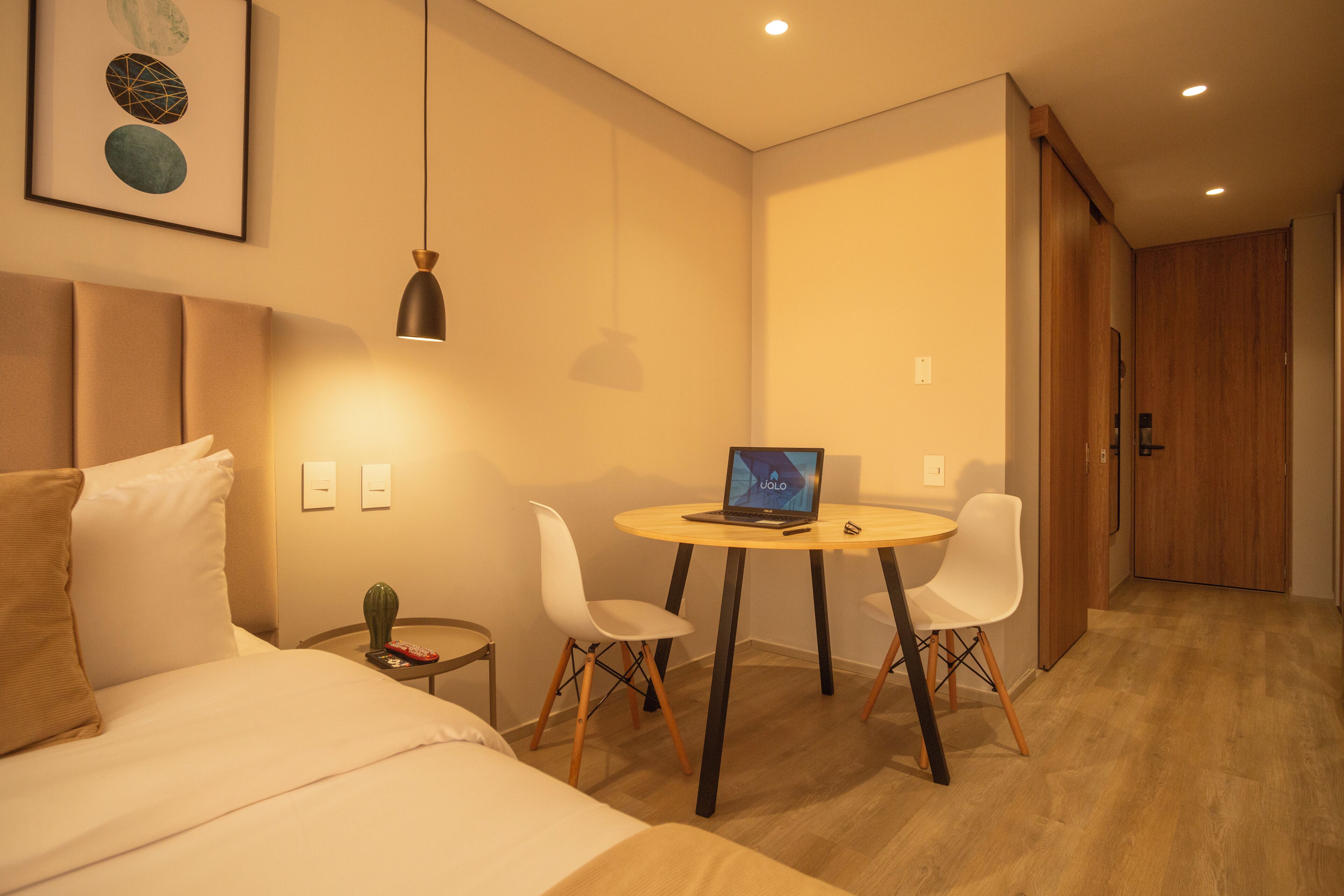 A modern hotel room with a bed, a desk with a laptop, two white chairs, a small side table, and a wooden door.