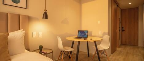 A modern hotel room with a bed, a desk with a laptop, two white chairs, a small side table, and a wooden door.
