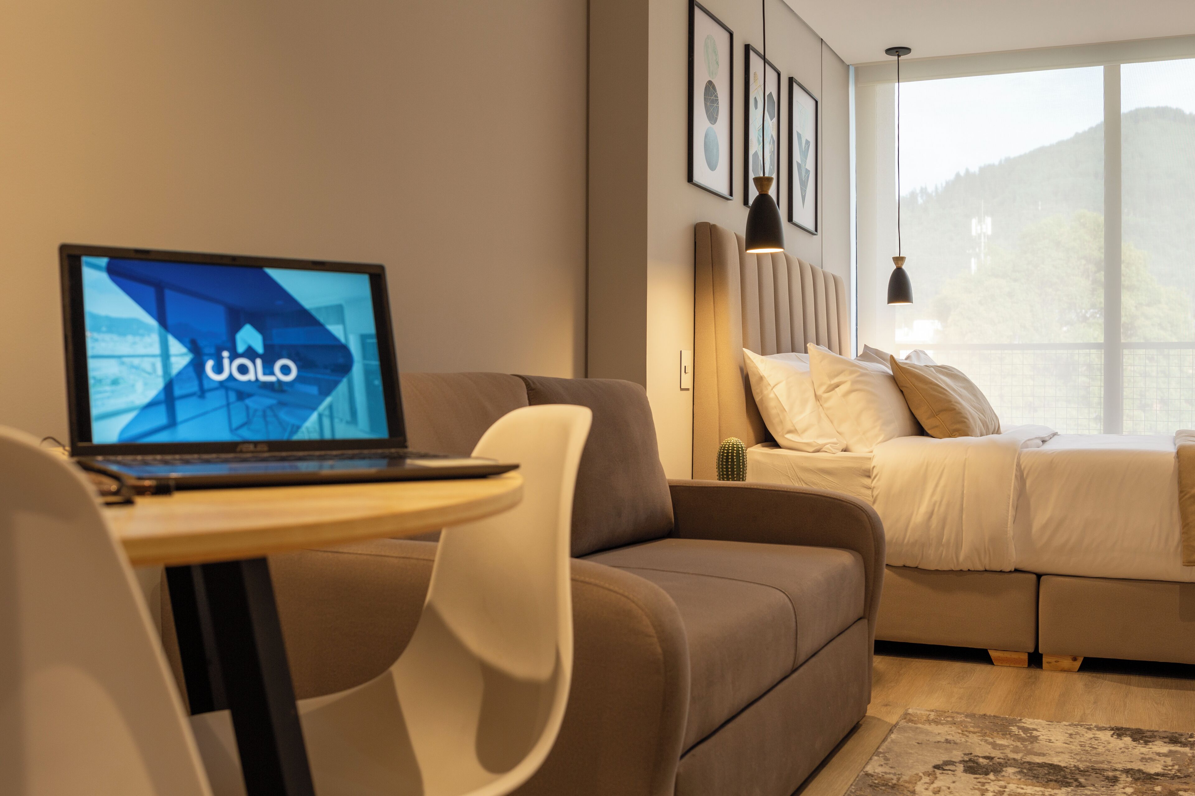 A modern hotel room with a bed, a sofa, a desk with a laptop, and a large window offering a view of the outdoors.