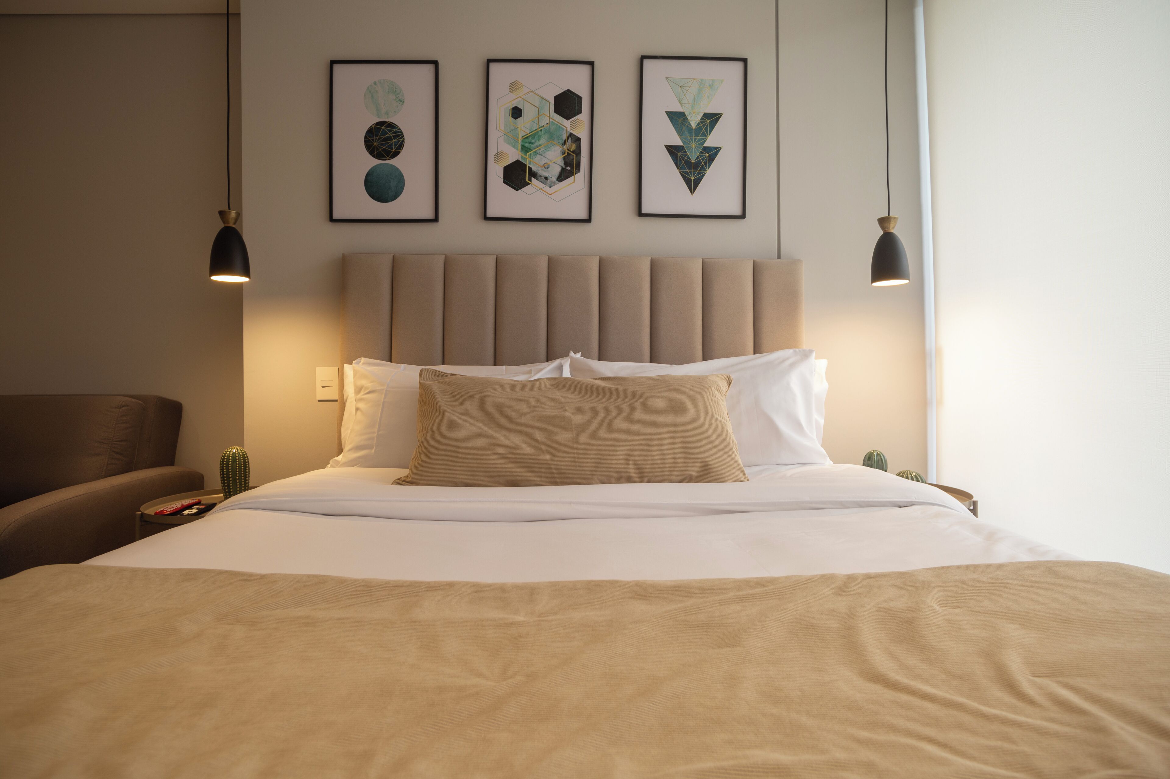 Un lit bien fait, avec une tête de lit beige, un fauteuil marron et trois œuvres d’art géométriques encadrées accrochées au mur.