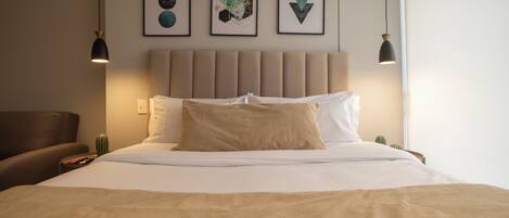 Un lit bien fait, avec une tête de lit beige, un fauteuil marron et trois œuvres d’art géométriques encadrées accrochées au mur.