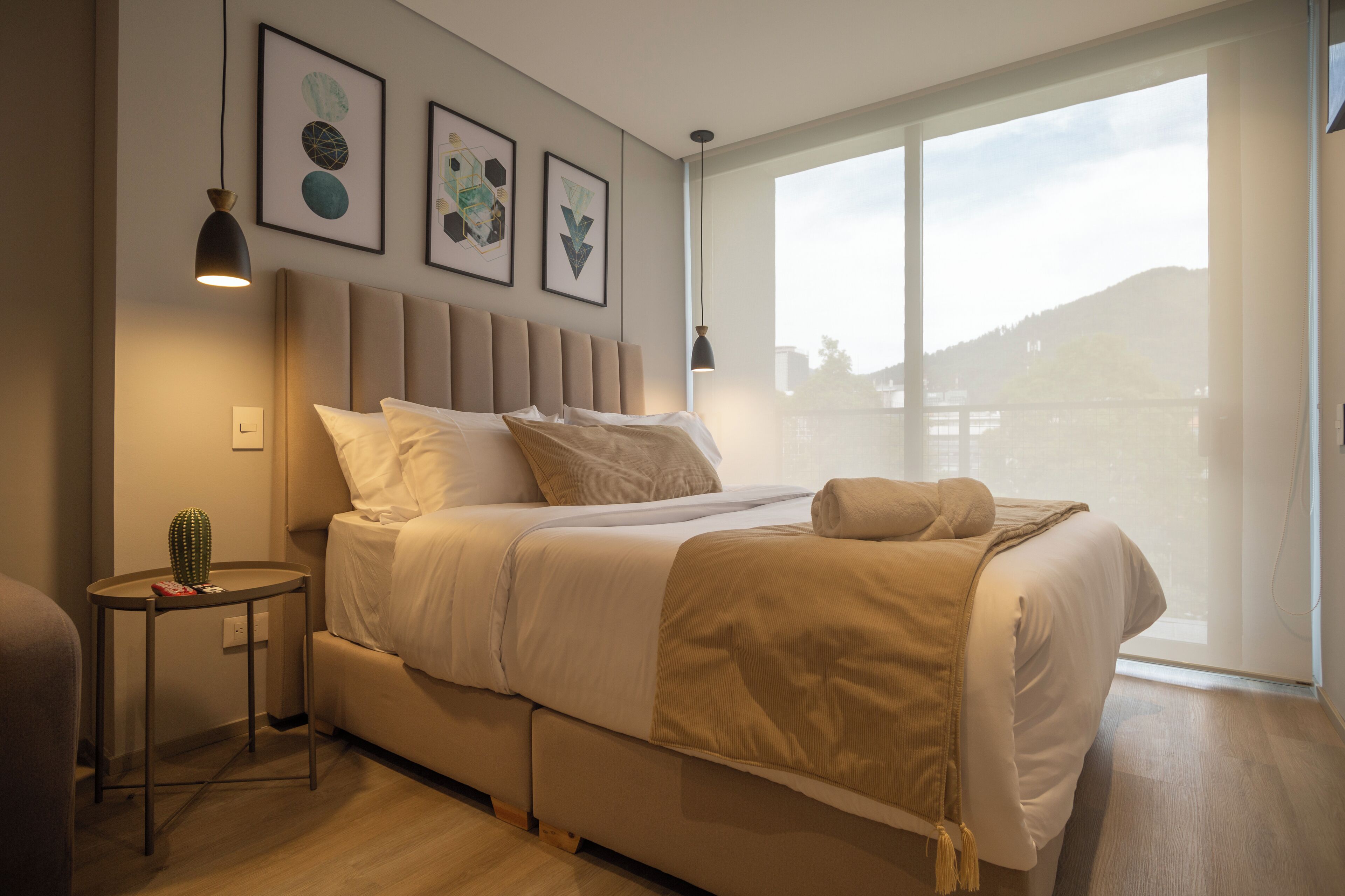 Moderni hotellihuone, jossa on suuri sänky, pieni sivupöytä ja ikkuna, josta avautuu näkymä kukkulalle.