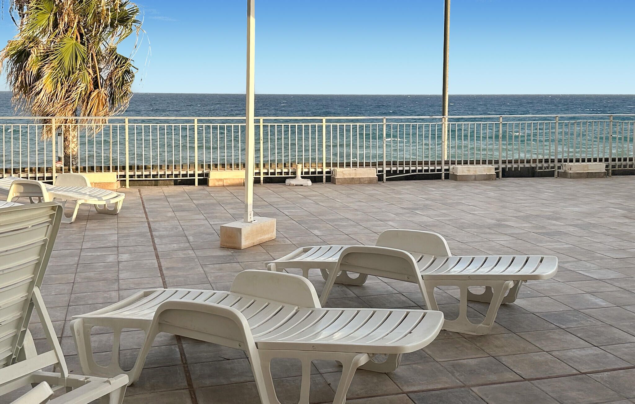 Un espacio al aire libre con sillas y mesas de plástico blanco, una barandilla de metal y vista al mar.