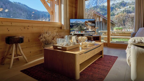 Ein Holzinnenraum mit einem großen Fenster, einem Fernseher, einem Holztisch und Blick auf Berge.