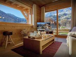 Interior de madera con un gran ventanal, un televisor, una mesa de centro de madera y vistas a montañas.