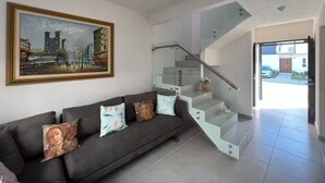 Een moderne woonkamer met een donkere bank, een glazen trap en een schilderij van een stadsgezicht.