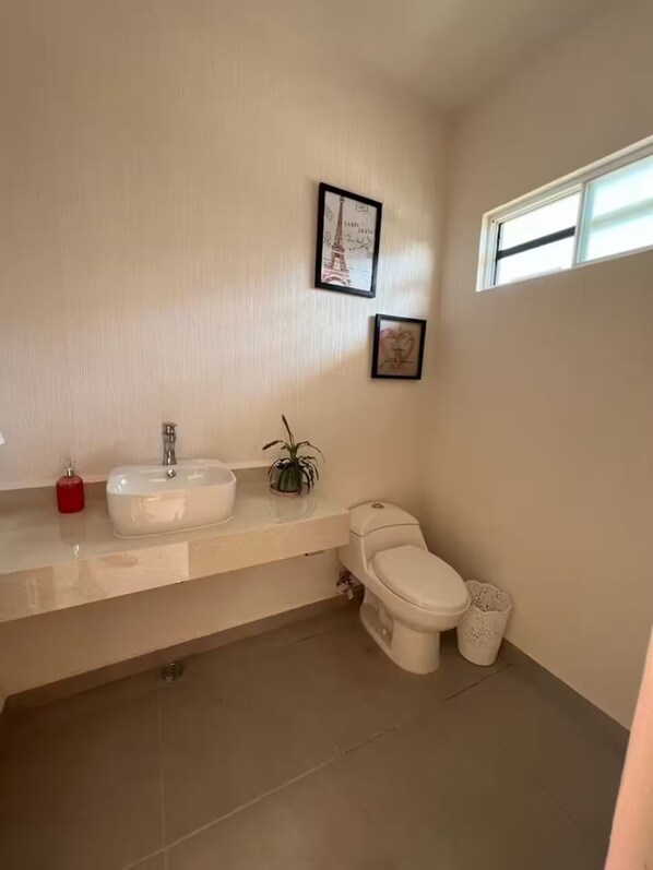 Un baño con un lavamanos blanco, inodoro y bidé, una ventana pequeña y dos cuadros en la pared.