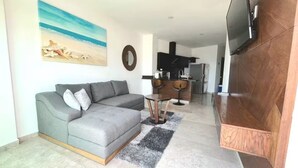 A modern living room with a grey sectional sofa, a small round coffee table, and a TV mounted on a wooden cabinet. A kitchen with dark cabinets and a refrigerator is visible in the background. - Depa Bucearías playa (Bucerías)