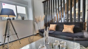 Een moderne woonkamer met een glazen salontafel, een donkere bank met kussens, een driepoot lamp en een groot raam met gordijnen.
