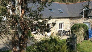 Une maison en pierre avec un toit en tuiles, une terrasse avec des meubles d’extérieur et un jardin agrémenté d’un arbre et de plantes.