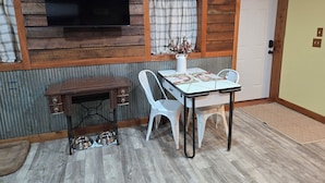 Une pièce avec un mur en bois, un téléviseur à écran plat fixé au-dessus d’une table recouverte d’une nappe blanche, deux chaises blanches, un petit bureau en bois avec des tiroirs et un bol pour chien en métal posé sur le sol.