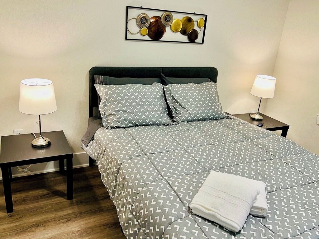 Ein ordentlich bezogenes Bett mit gemusterten Betttextilien, zwei Nachttischlampen und ein an der Wand befestigtes abstraktes Kunstwerk.