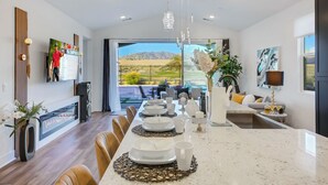 A modern kitchen with a long dining table set for six, a TV mounted on the wall, and a view of a tennis court and mountains through large windows.