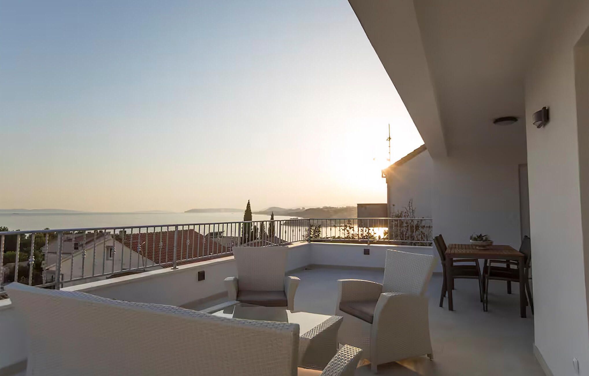 A balcony with wicker furniture, a table, and a view of the sea and distant land during sunset.