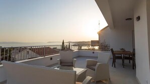 A balcony with wicker furniture, a table, and a view of the sea and distant land during sunset. - Lovely apartment in Podstrana with WiFi (Podstrana)