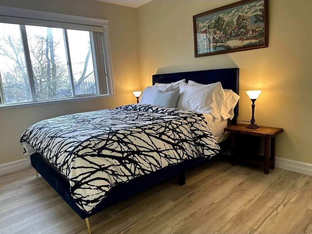 Een slaapkamer met een groot bed, twee bedlampjes, een schilderij aan de muur en een raam met uitzicht op bomen.