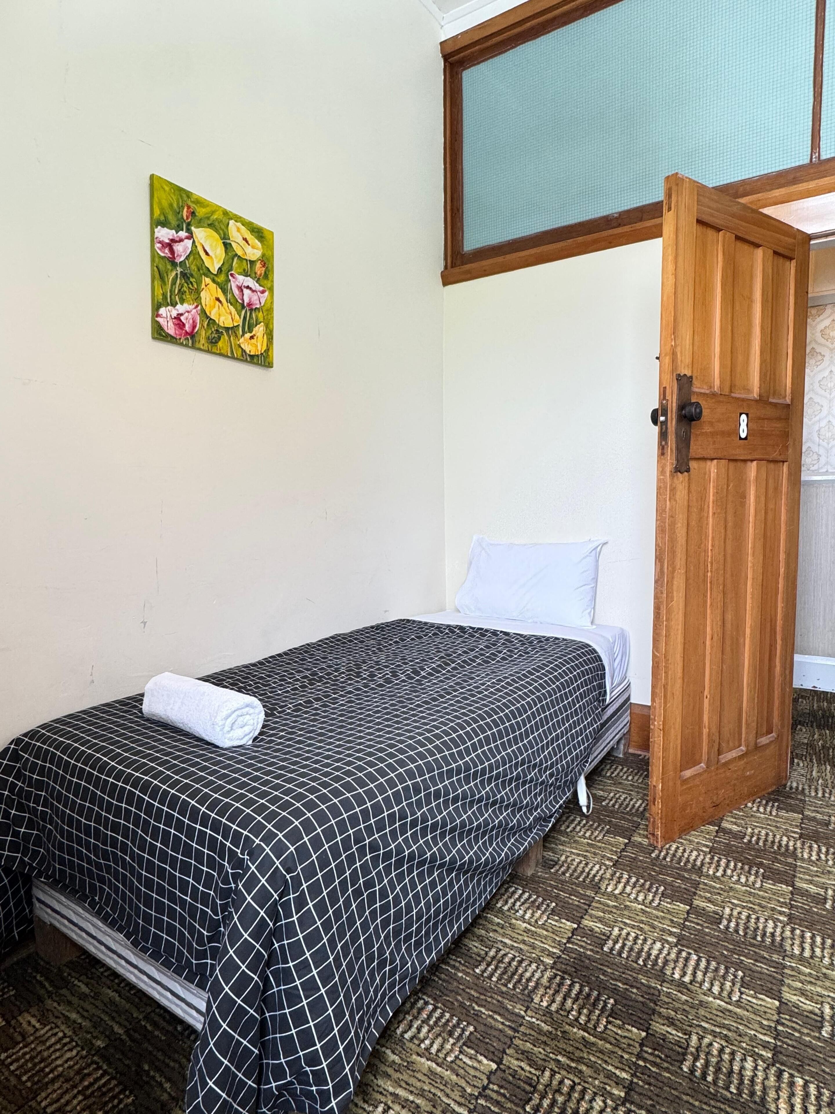 A single bed with a black and white checkered bedspread, a rolled white towel, a green floral painting on the wall, and a wooden door with a number plate.