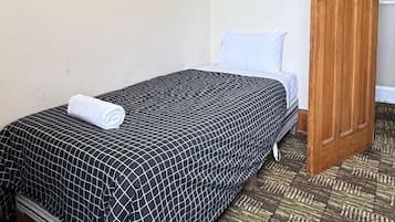 A single bed with a black and white checkered bedspread, a rolled white towel, a green floral painting on the wall, and a wooden door with a number plate.
