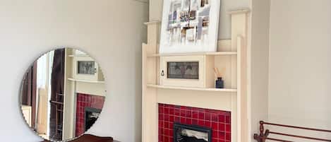 A room with a wooden dresser, a mirror, a fireplace with red tiles, and a framed artwork above the fireplace.