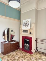Quarto com cômoda de madeira, um espelho, lareira com azulejos vermelhos e uma obra de arte emoldurada acima da lareira.