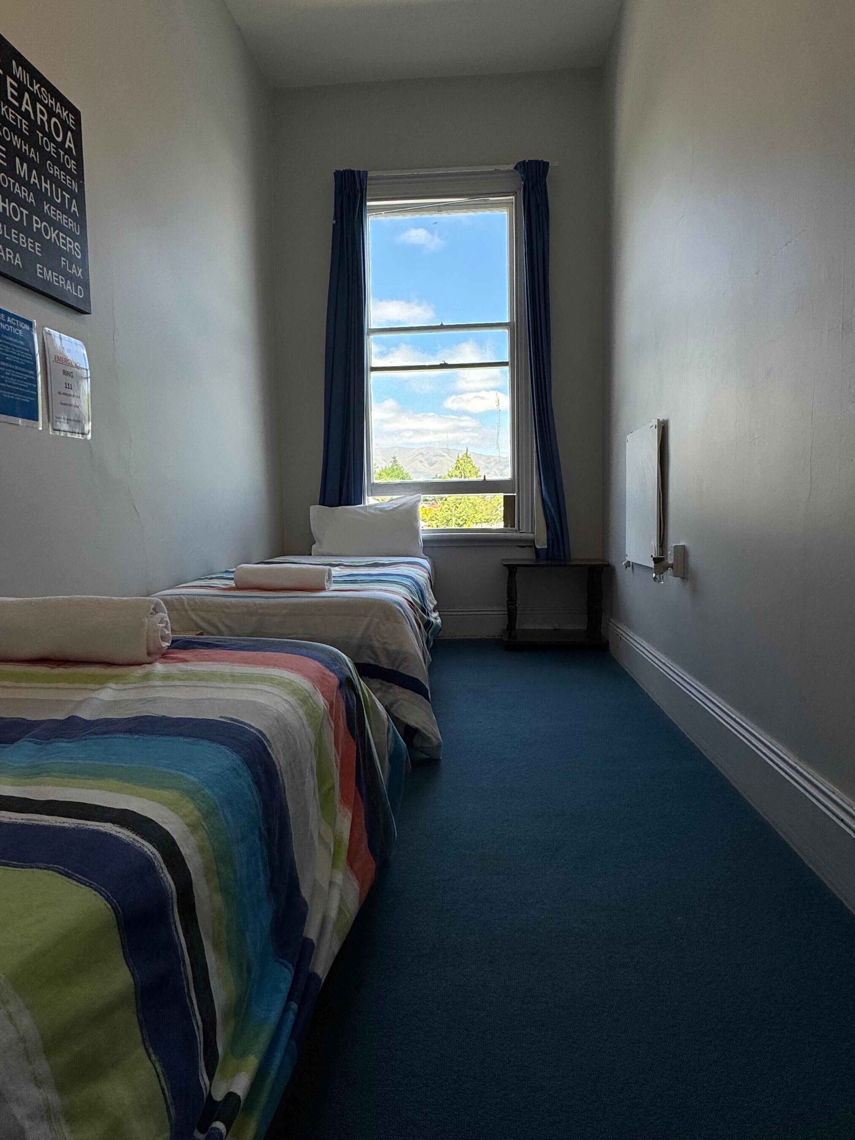 A narrow hotel room with two beds, a window with curtains, a radiator, and a view of the outdoors.