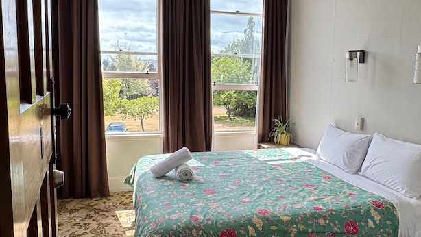 Habitación de hotel con una cama grande, colcha con motivos florales y vistas al exterior con vegetación, además de una ventana con cortinas.
