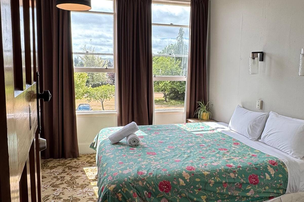 Habitación de hotel con una cama grande, colcha con motivos florales y vistas al exterior con vegetación, además de una ventana con cortinas.