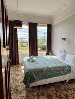 Quarto de hotel com uma cama grande, colcha florida e vista para o verde lá fora, com uma janela e cortinas.