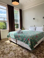 Uma cama bem arrumada com colcha floral, uma janela com cortinas e um aplique na parede.