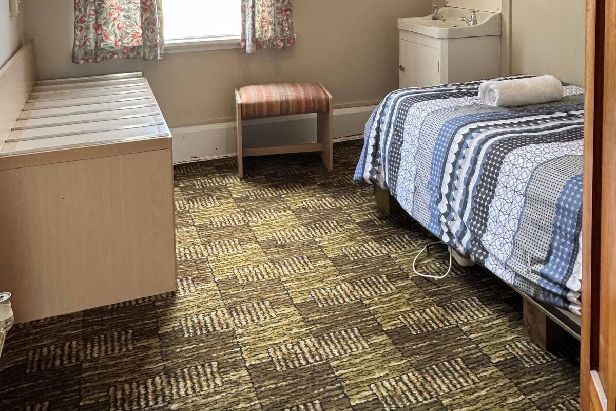 Un dormitorio con cama, mesita de noche, un taburete pequeño, ventana con cortinas, lámpara colgante, espejo y lavamanos.