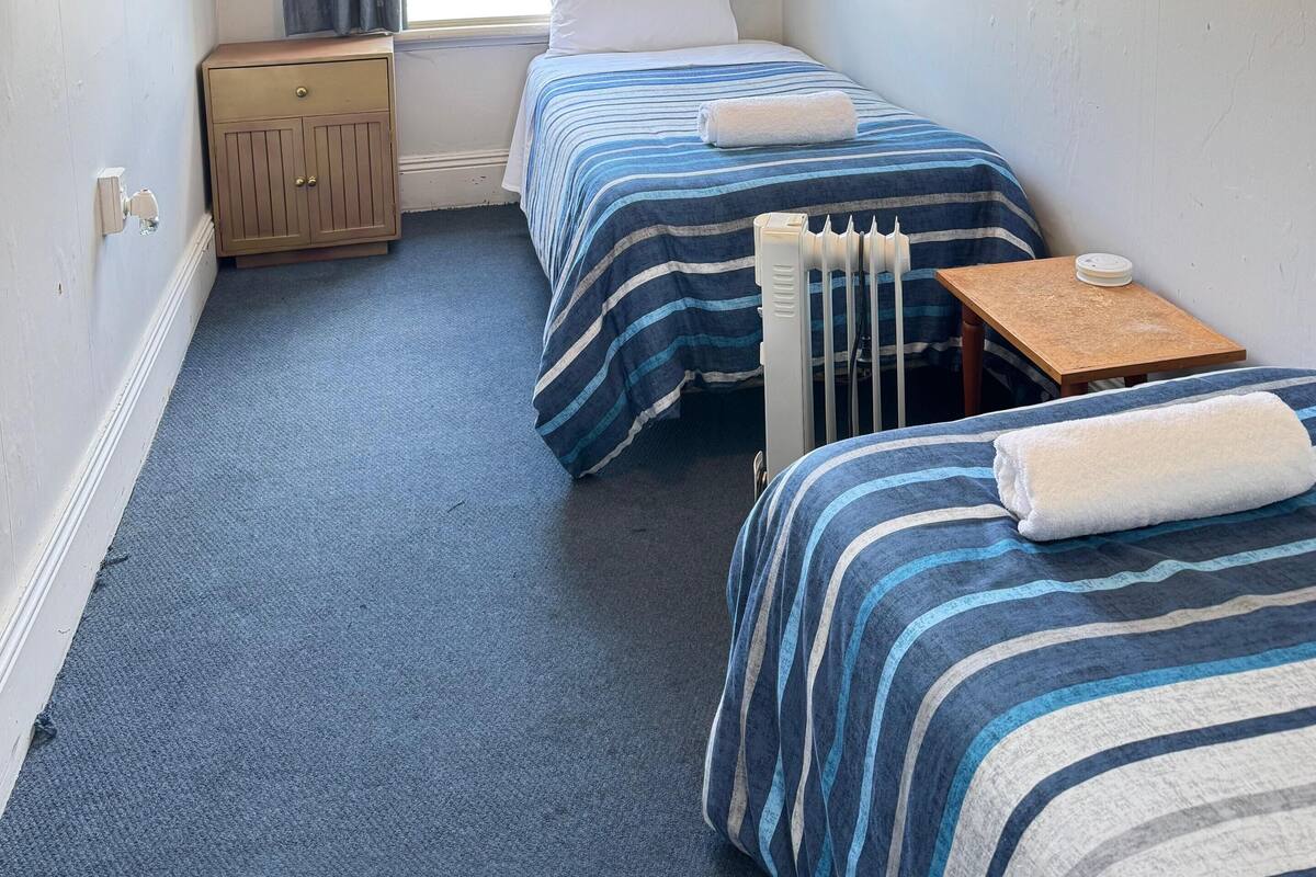 Una habitación pequeña y bien iluminada con dos camas, un armario de madera, un radiador y una ventana con cortinas.