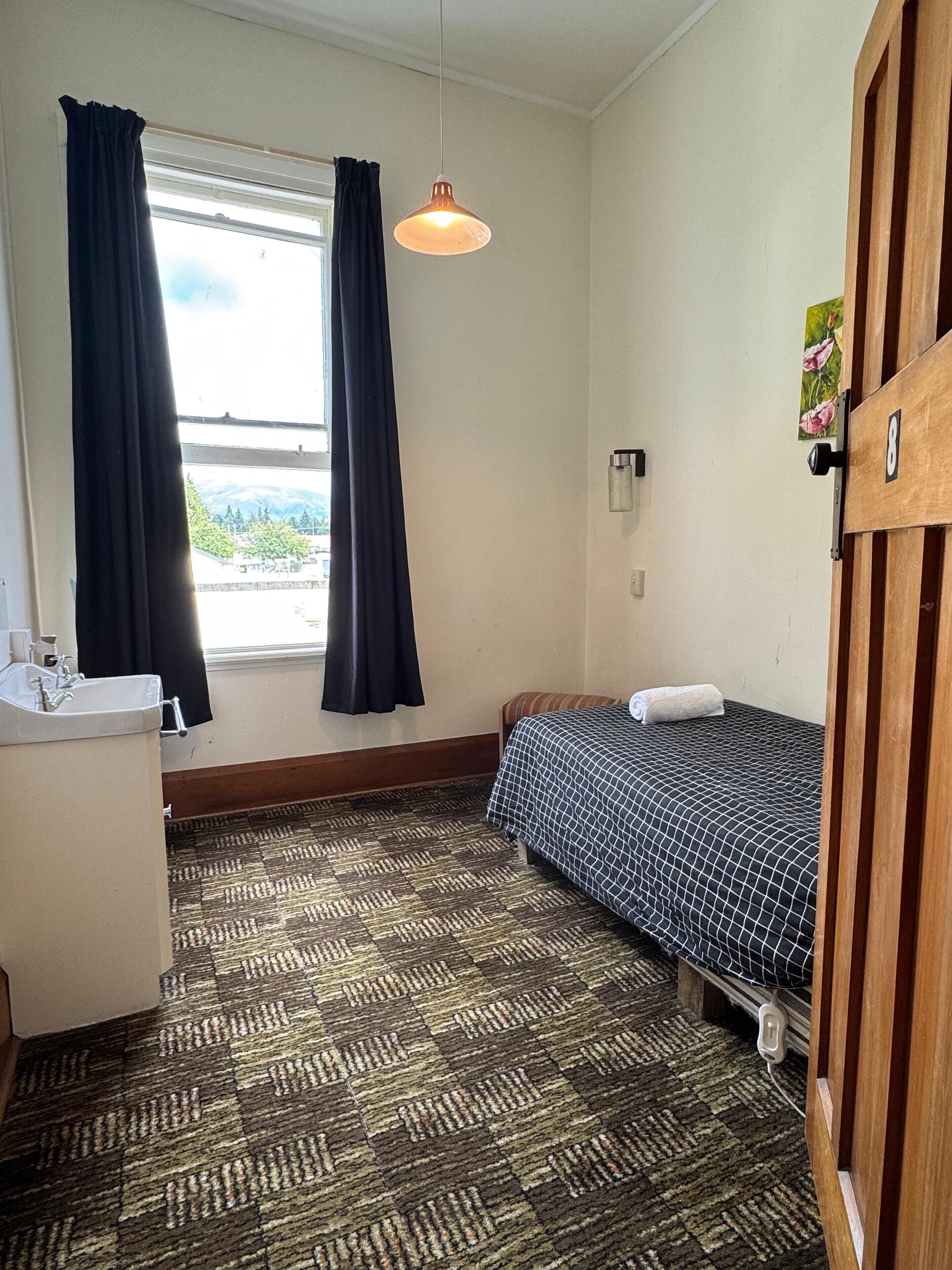 A small, well-lit room with a bed, a window with curtains, a hanging light fixture, and a wooden door.