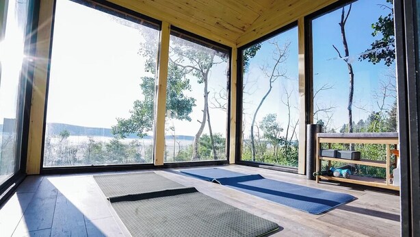 A room with large windows, wooden flooring, and exercise mats.
