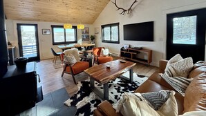 A modern living room with a wooden ceiling, a large TV, a dining area with a table and chairs, and a cozy seating area with a leather couch and a cowhide rug.