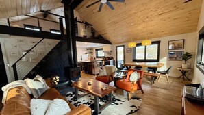 A spacious living area with a wooden ceiling, a fireplace, a dining table, and a kitchen in the background.