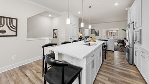 Una cocina moderna con armarios blancos, una isla central con taburetes y luces colgantes, y vista al área de estar.