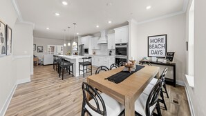 Una cocina moderna con armarios blancos, una mesa de comedor de madera y sillas negras. En la pared hay un cartel que dice "More Beach Days".