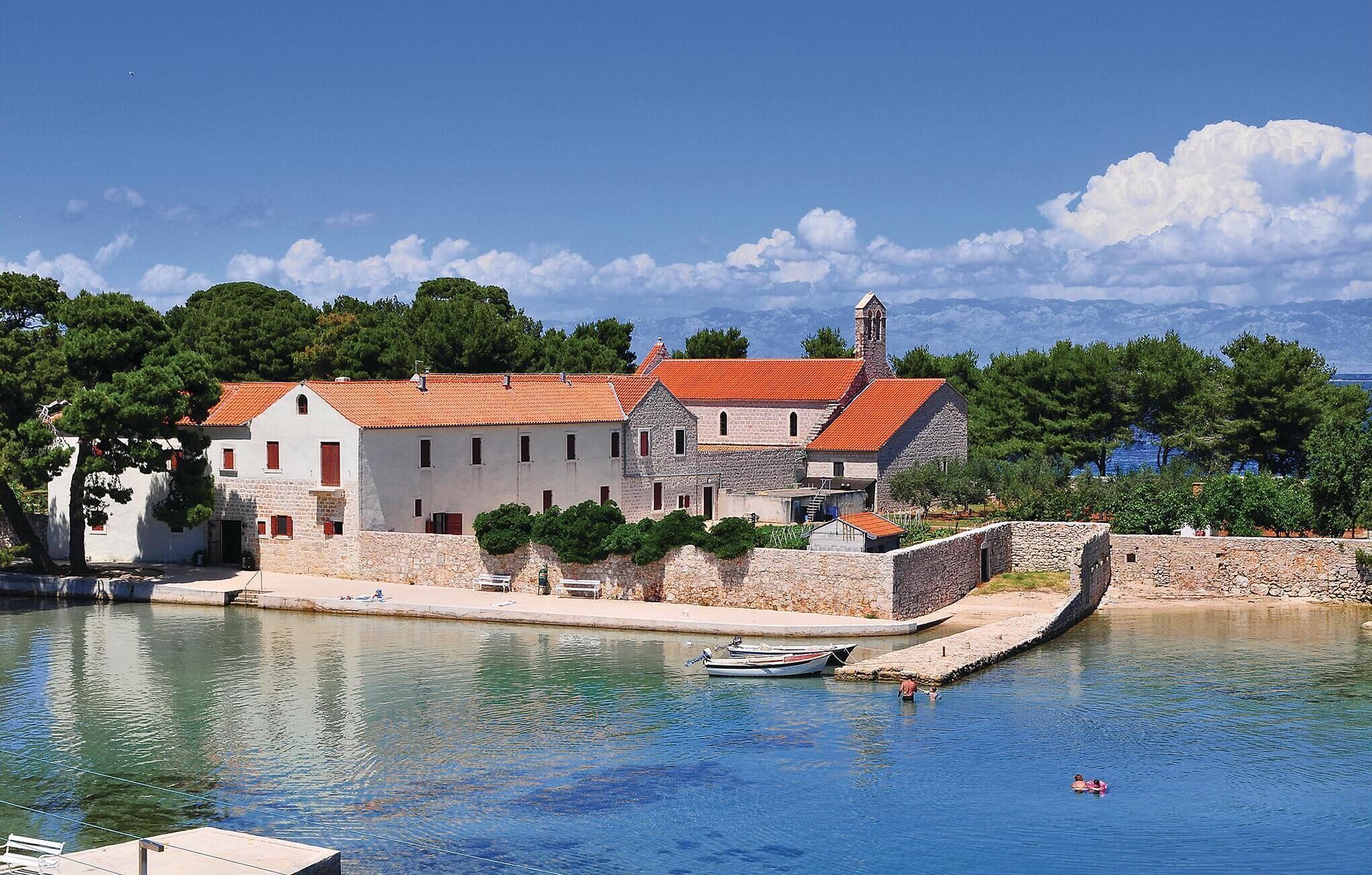 Un bâtiment historique à la façade en pierre et au toit recouvert de tuiles rouges, situé au bord d’une étendue d’eau calme, avec des bateaux amarrés à proximité.
