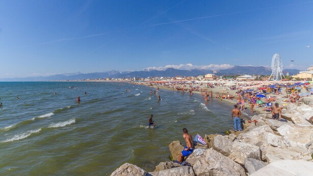 Ein belebter Strand mit badenden und sonnenden Menschen, einem Riesenrad und Bergen im Hintergrund.