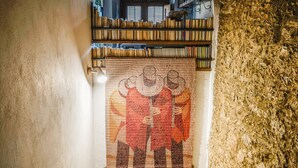 Ein Zimmer mit einem großen Wandteppich, der eine historische Szene zeigt, einem Bücherregal voller Bücher und einer strukturierten Steinwand.