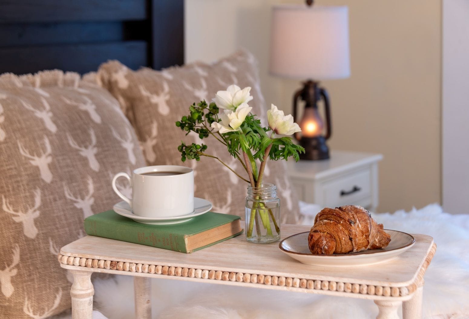 Un dormitorio acogedor con una mesita de noche que sostiene una taza de café, un libro, un florero con flores y un plato con un croissant.