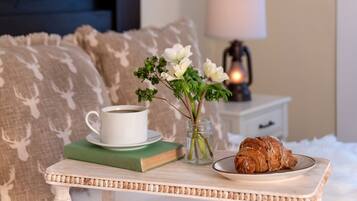 Un dormitorio acogedor con una mesita de noche que sostiene una taza de café, un libro, un florero con flores y un plato con un croissant.