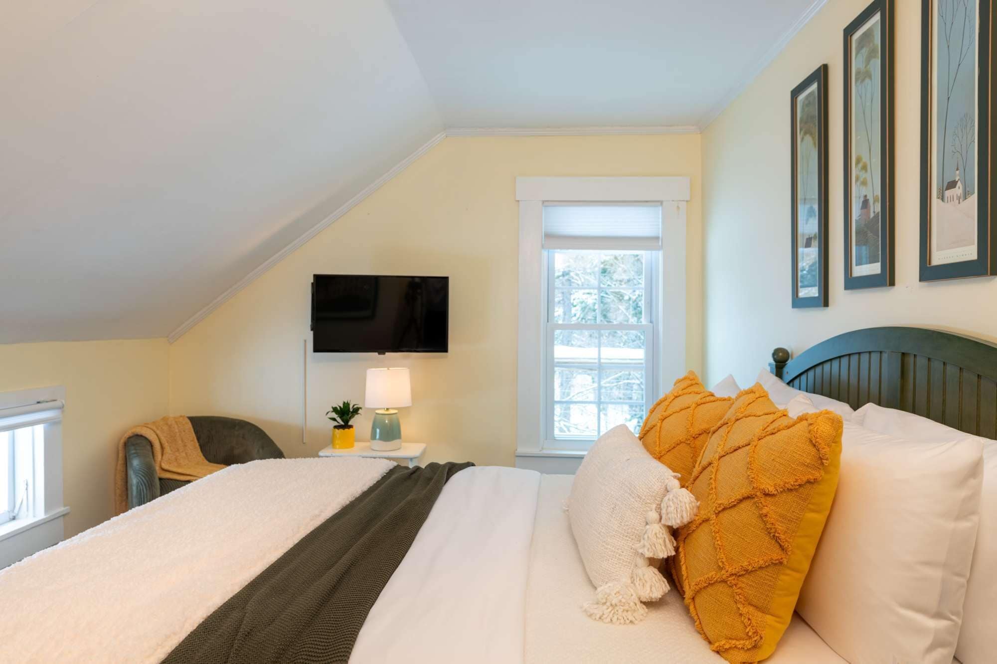 A bedroom with a bed, a TV mounted on the wall, a window with a view of trees, and framed pictures on the wall.