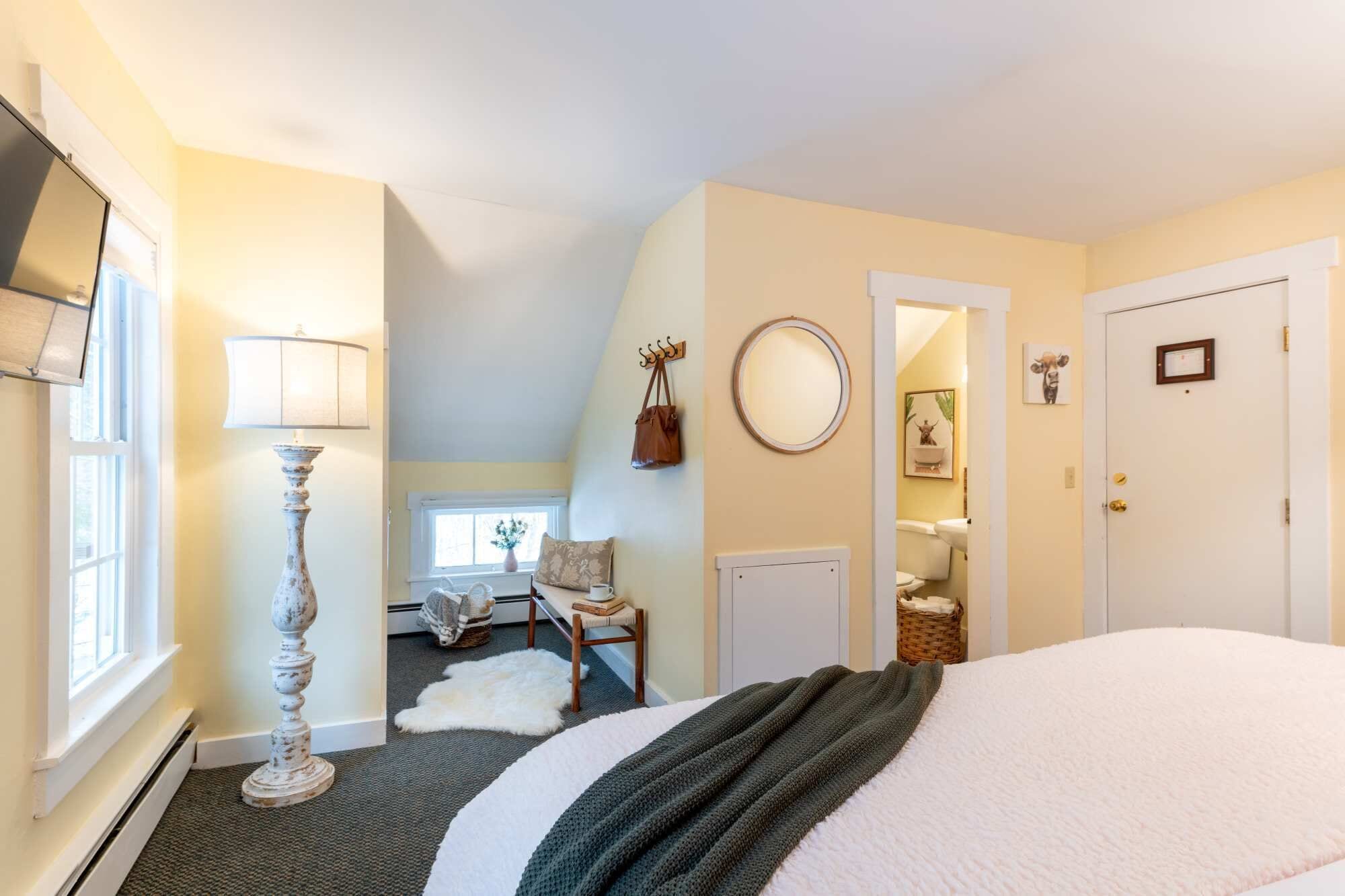 A bedroom with a large bed, a lamp, a chair, a small table, a mirror, and a door.