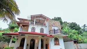 Une maison à deux étages, avec un toit recouvert de tuiles rouges, un balcon et un jardin agrémenté de plantes en pot.