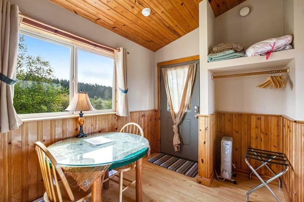 Une pièce chaleureuse avec un plafond en bois, une table en verre, des chaises, une lampe, une porte avec des rideaux, un radiateur et une étagère avec des serviettes pliées.