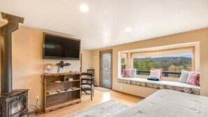 A bedroom with a large bed, a TV, a fireplace, a window seat with pillows, and a view of the outdoors. - Sunset Suite in Deer Harbor (Eastsound)