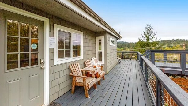 A wooden deck with Adirondack chairs and a table, a covered porch area, and a view of a forested landscape. - Sunset Suite in Deer Harbor (Eastsound)